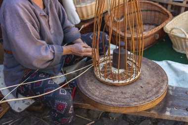 One elderly unrecognizable caucasian woman in old ancient clothes sits on a bench at a small table and weaves a basket of twigs pressed down by a weight, side view, close-up. Concept of medieval needlework, artisanal.