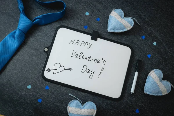 One empty marker board with the inscription: happy valentine's day, blue tie, three textile hearts and shiny confetti lies diagonally against a black stone background, flat lay close-up. Valentine's day concept.