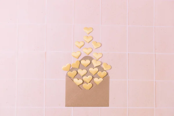 One craft envelope with yellow decorative hearts flowing from it lies in the center on a pale pink tiled background with copy space on the sides, flat lay close-up. Valentine\'s day concept, love message.