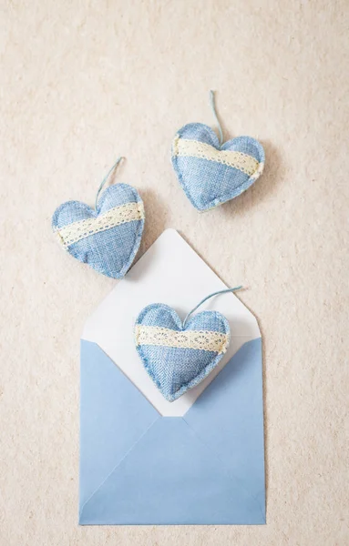 One envelope with three drop-down textile hearts with openwork stripes on the left against a natural beige stone background with copy space on the right, flat close-up. Valentine's day concept.