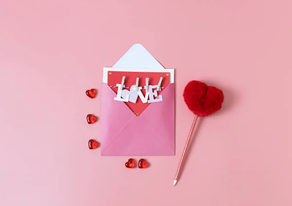 Open envelope with letter paper word love, red glass hearts and a pen lie in the center of a pink background, flat close-up. Valentine\'s day concept.