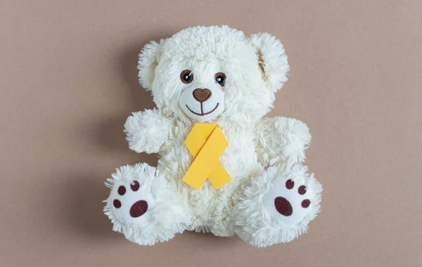 One toy white bear with yellow paper ribbon tie lies in the center on a natural nude background, flat lay close-up. World childhood cancer day concept.