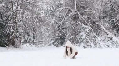 Beyaz ceketli, kahverengi pantolonlu ve örgü eldivenli güzel beyaz esmer bir kız kış ormanındaki bir açıklıkta dikilirken kar fırlatıyor. Kış yürüyüşü, kış eğlencesi.