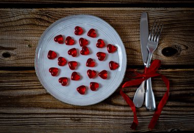 Beyaz bir tabakta kırmızı cam kalpler, çatal ve kırmızı kurdeleye sarılı bir bıçak kahverengi tahta bir masanın ortasında, düz bir zeminde yatıyor. Sevgililer Günü konsepti, şenlik sofrası kurma, şenlik masası kurma..