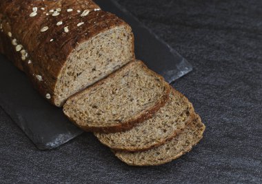 One rye dark bread with crushed oat grains and sunflower seeds lies cut into thin pieces lies on a stone stand on the left on a black background, flat lay close-up.The concept of baking bread.