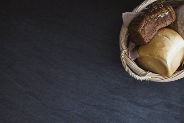 Three different varieties and shapes in an oval wicker basket with handles lie to the right on a black stone table with copy space on the left, flat lay close-up. The concept of baking bread.