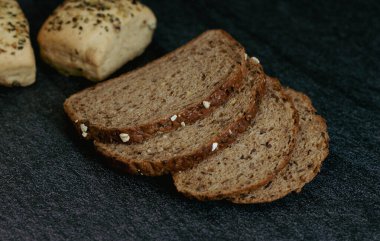 Four slices of rye bread with fiber and two buns with seeds lie on a black stone table with a blurred background, close-up top view. The concept of baking bread.