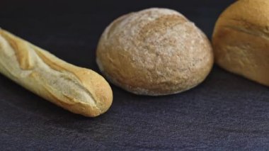 Two breads of different varieties with one French baguette lie against a background of black stone with tracking to the right, close-up side view. The concept of baking bread.