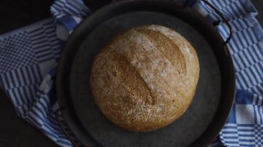One round rye bread in a vintage metal dish with a blue checkered kitchen napkin lies in the center on a black stone background, top view close-up tracking to the right. The concept of baking bread.