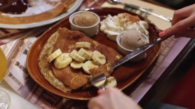 A young Caucasian unrecognizable girl pricks a sweet pancake with a banana on a fork and eats it while sitting at a table in a restaurant, close-up side view. Women lifestyle concept.