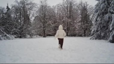 A young caucasian girl in a white down jacket with a hood and brown pants from the back runs in the winter forest of the reserve in belgium, close-up side view. Winter holiday concept, modern lifestyle.