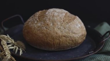 One round rye bread in a vintage metal platter with ears and an olive kitchen napkin lie on a black stone background, zoom side view close-up. The concept of baking bread.