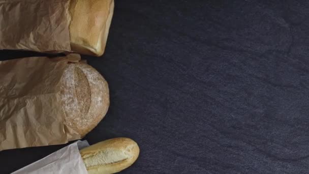 两个不同的面包和一个纸袋中的法式面包放在右边的一块黑色石头背景上 左边有复制空间 并且追踪到右边的 关闭的侧面视图 烘焙面包的概念 — 图库视频影像