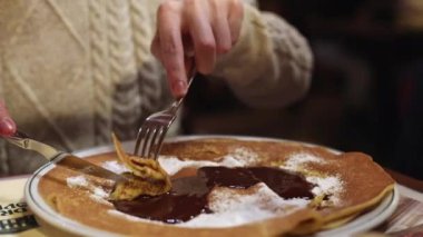 A young Caucasian unrecognizable man dunks a sweet pancake in melted chocolate with a fork and eats it while sitting at a table in a restaurant, close-up side view. Men's lifestyle concept.