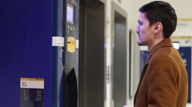 A young caucasian handsome brunette man in a brown coat makes a payment in an underground parking terminal, close-up side view. Men's lifestyle concept, using technology.