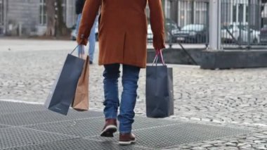 Kahverengi ceketli, elinde alışveriş poşetleri olan genç ve şık bir beyaz adam meydanın taş kaldırım taşları boyunca yürür metronun yeraltı geçidine kadar, çevrimdışı alışveriş konsepti, modern yaşam tarzı.