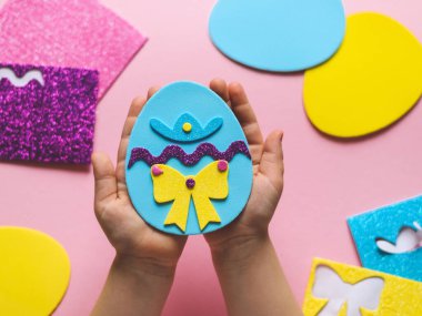 The hands of a little caucasian girl show the end result of a homemade felt easter egg, holding in her palms and sitting at a children's table with a set of crafts on a pink background with depth of field, flat lay close-up. The concept of crafts, di