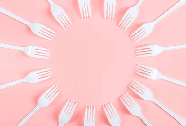 White plastic forks are arranged in a round frame on a light pink background with copy space for your text in the center, flat lay close-up. The concept of ecology and plastic garbage and disposable plastic tableware.