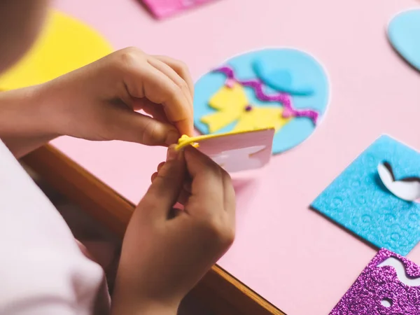 The hands of a little caucasian girl peel off a yellow felt sticker for a blue egg with her fingers, sitting at a children\'s table with a set of craft preds on a pink background with depth of field, close-up side view. The concept of crafts, diy, nee