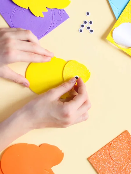 The hand of a caucasian teenage girl glues a decorative eye on a felt yellow chick, sitting at a table with a set of craft objects on a pale yellow background, close-up flat lay. The concept of diy, crafts, needlework, handicraft, children\'s art, at