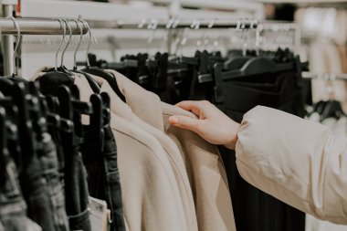 The hand of a young unrecognizable caucasian girl chooses a light beige women's coat hanging on a hanger between black jeans on a stand in a clothing boutique, shopping, close-up side view.