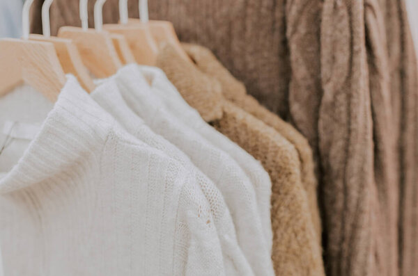 White knitted turtleneck sweaters and brown cardigans hang on hangers on a stand in a clothing store, close-up side view with selective focus.Offline shopping concept.