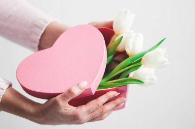 The hands of a young caucasian woman with a beautiful and delicate manicure hold a pink heart box against the background of a white wall, side view close-up, depth of field. Valentines day concept