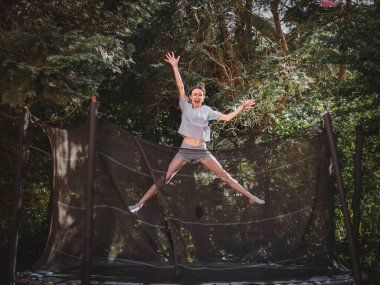 A beautiful slender caucasian teenage girl in shorts and a blouse jumps on a trampoline, frozen in a funny position of a star in the garden of a house among trees, close-up side view. Concept at home, home sports, happy childhood, children entertainm