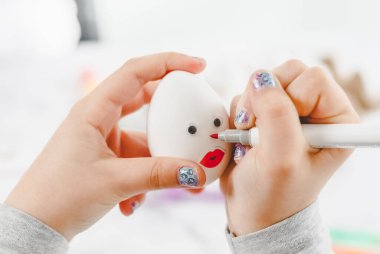 Children's hands hold one white egg with glued eyes, lips and draw a nose with a marker, sitting at a marble table with depth of field, preparing diy for the Easter holiday, close-up side view. The concept of crafts, artisanal, home, children's creat