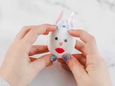 Children's hands hold one white egg with glued eyes, lips, bow tie and glue paper bunny ears, sit at a marble table with depth of field, prepare crafts for the Easter holiday, close-up side view. The concept of craft, needlework, diy, children's crea