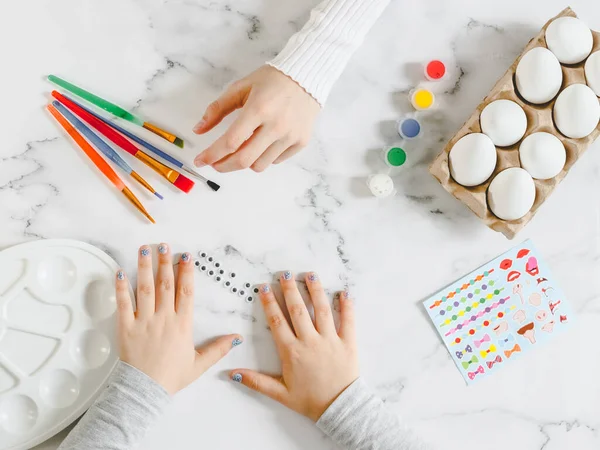 Two pairs of children\'s hands are outstretched to the brushes and glued eyes on a marble table with white eggs in a cardboard box, acrylic paint, stickers and a palette for diy preparation for the Easter holiday, close-up flat lay. The concept of cra