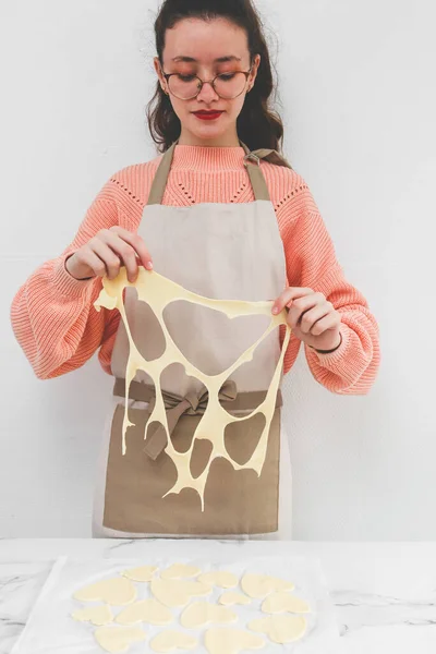 A young caucasian teen baker girl in an apron separates raw dough from cut out hearts on a white parchment at a marble table against a marble wall, close-up side view. Cooking at home, homemade cookies and valentine's day concept.
