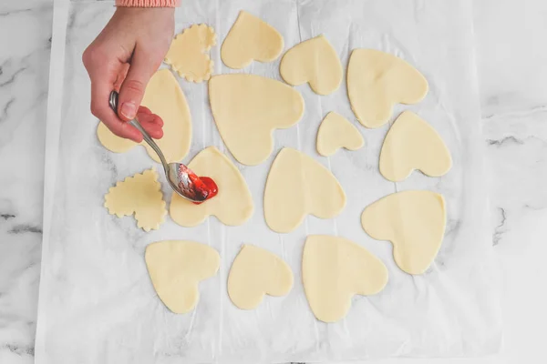 The hands of a young caucasian baker girl are smeared with strawberry jam cut out hearts of various shapes and sizes from raw dough on a white parchment at a marble table, close-up side view. Cooking at home, homemade cookies and valentine\'s day conc