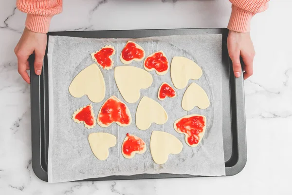 The hands of a young caucasian baker girl hold a baking sheet with heart cookies smeared with strawberry jam from raw dough on a white parchment at a marble table, flat lay close-up. Cooking at home, homemade cookies and valentine's day concept.