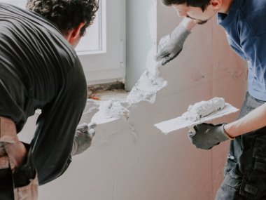 Young caucasian guys plasterers in blue and gray t-shirts apply putty with spatulas on the wall from below the window, close-up side view. The concept of apartment and house renovation, wall