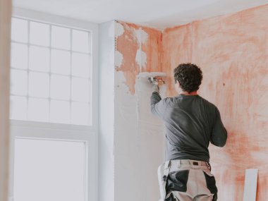 A young caucasian guy in a gray t-shirt with sleeves, work pants and curly brown hair applies fresh putty with a small spatula on the wall near the window, standing on a stepladder, close-up side view