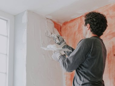 A young caucasian guy in a gray t-shirt with sleeves and curly brown hair applies fresh putty with a small spatula on the wall near the window while standing on a stepladder, close-up side view. The