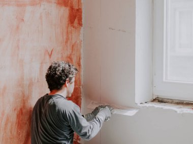 A young caucasian plasterer in gray long sleeve t-shirts, brown curly hair and construction gloves from the back holds a large trowel and levels a freshly plastered wall while squatting on the floor