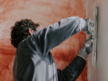 A young caucasian guy in a gray t-shirt with sleeves and curly brown hair applies fresh putty with a large spatula on the wall, close-up side view. The concept of apartment and house renovation, wall