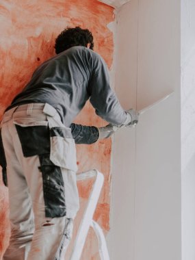 A caucasian young guy, a plasterer in a gray T-shirt with sleeves, work pants and curly brown hair, smoothes fresh putty with a large spatula on the upper wall near the ceiling, pulling down and