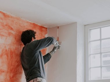 A young caucasian guy in a gray t-shirt with sleeves and curly brown hair removes a construction beacon with a spatula after he puttied the wall near the window while standing on a stepladder, close