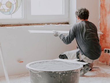 A young caucasian plasterer in uniform with brown curly hair plasters the walls and checks them for evenness with a large wooden rule, squatting from the back, next to him is a black basin with fresh
