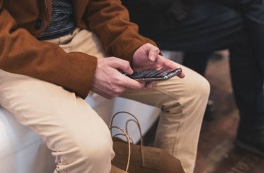 One young caucasian guy in a brown coat and beige trousers reads an article in a smartphone while sitting on a plastic white bench in a store in a mall, close-up top view. The concept of using technology, online shopping.