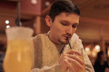 Portrait of one young handsome brunette caucasian man in knitted sweater drinking a cocktail with a straw from a glass, sitting at a table in a restaurant, close-up side view. Men lifestyle concept, real emotions.