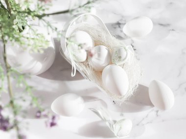 Decorative white eggs, a metal egg basket and a ceramic vase with spring flowers on a marble table in the early morning, flat lay close-up. Happy easter concept, holiday banner.