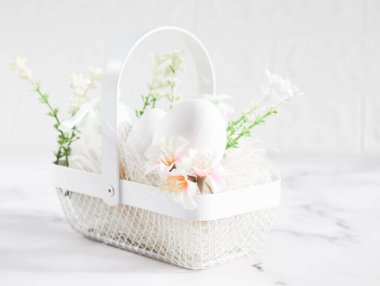 Decorative white eggs in a metal basket with eggs, spring flowers and straw stand on the left on a marble table in the early morning, close-up side view. Happy easter concept, holiday banner.