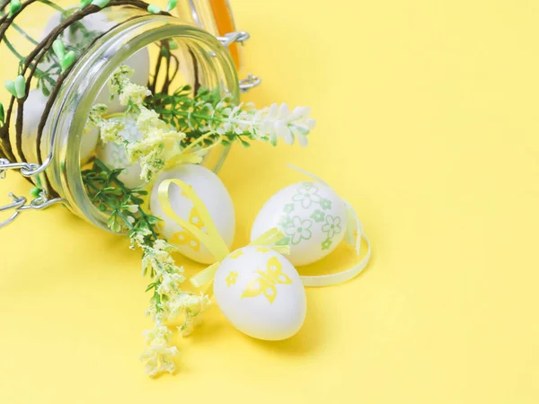 Decorative Easter eggs and spring flower scattered from a lying glass jar on a yellow background, close-up side view. Happy easter concept, holiday banner.