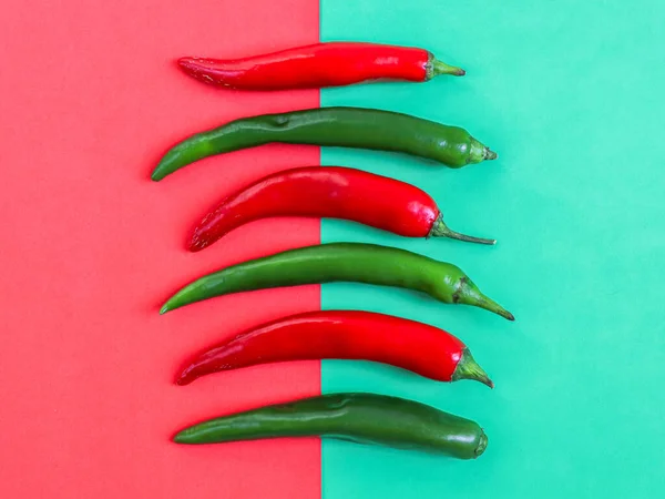 Six hot green and red chili peppers lie vertically in a row on a red-green background, flat lay close-up. The concept of Cinco de Mayo, minimalism, blanks.