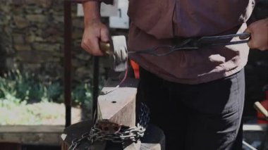 Middle-aged Caucasian male blacksmith of the medieval era forges red-hot iron by pounding on it with a sledgehammer, then transfers it to the fire, standing in the forge on a summer day,side view, slow motion. Artisanal concept, medieval lifestyle