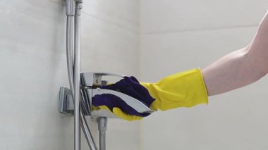 The hands of a caucasian young woman in yellow rub a dark lilac microfiber cloth over a wet faucet in the bathroom, side view close-up in slow motion.Cleaning the bathroom concept.